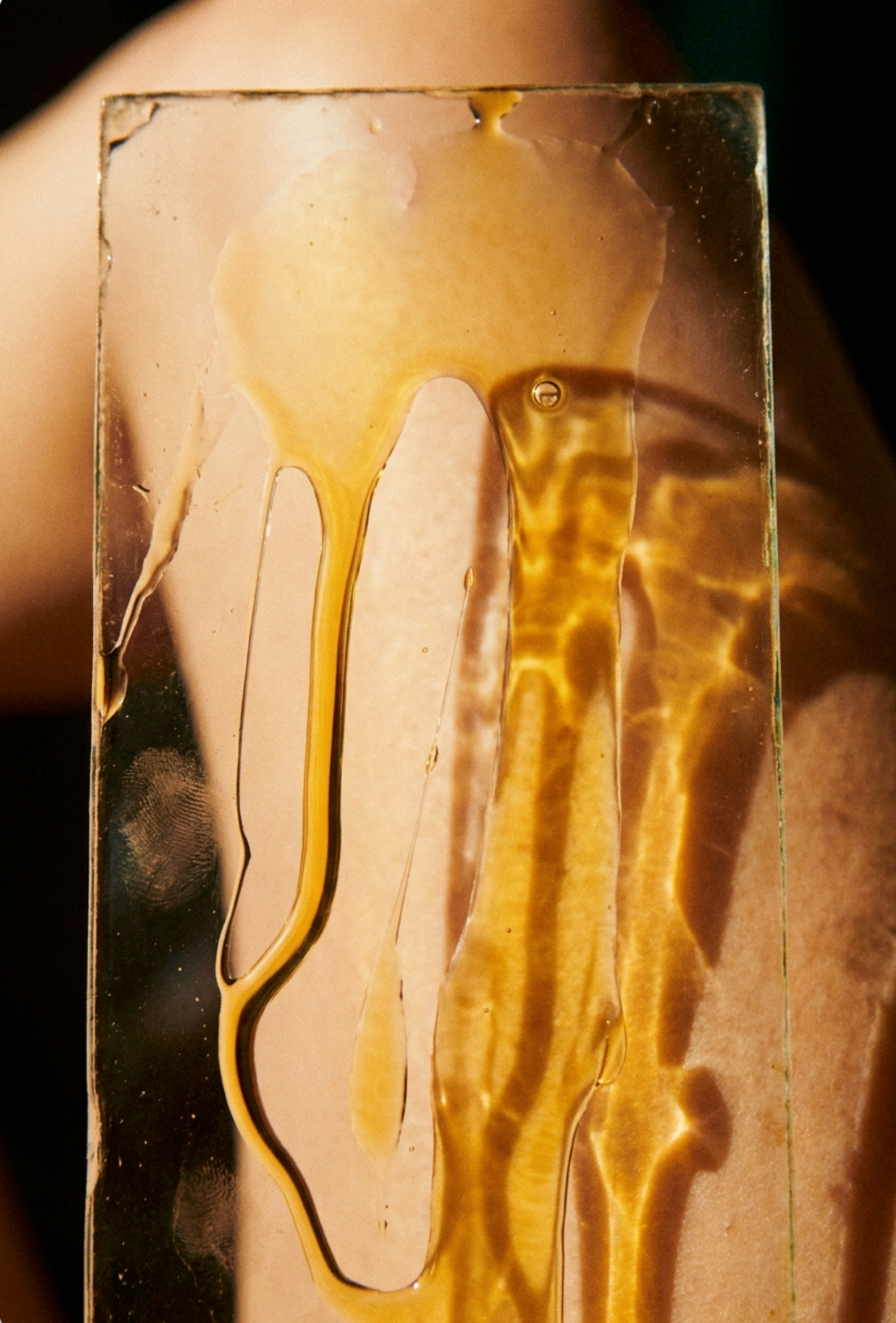 Golden face oil running down a pane of glass held against skin