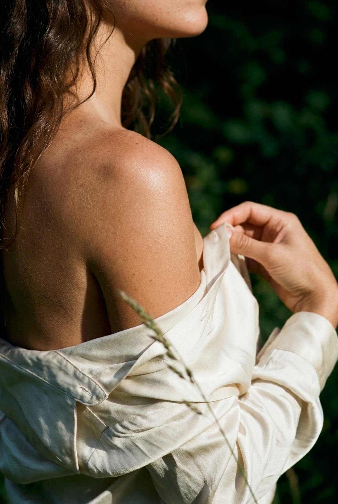 Woman outdoors in greenery slipping a cream shirt off her bare shoulder