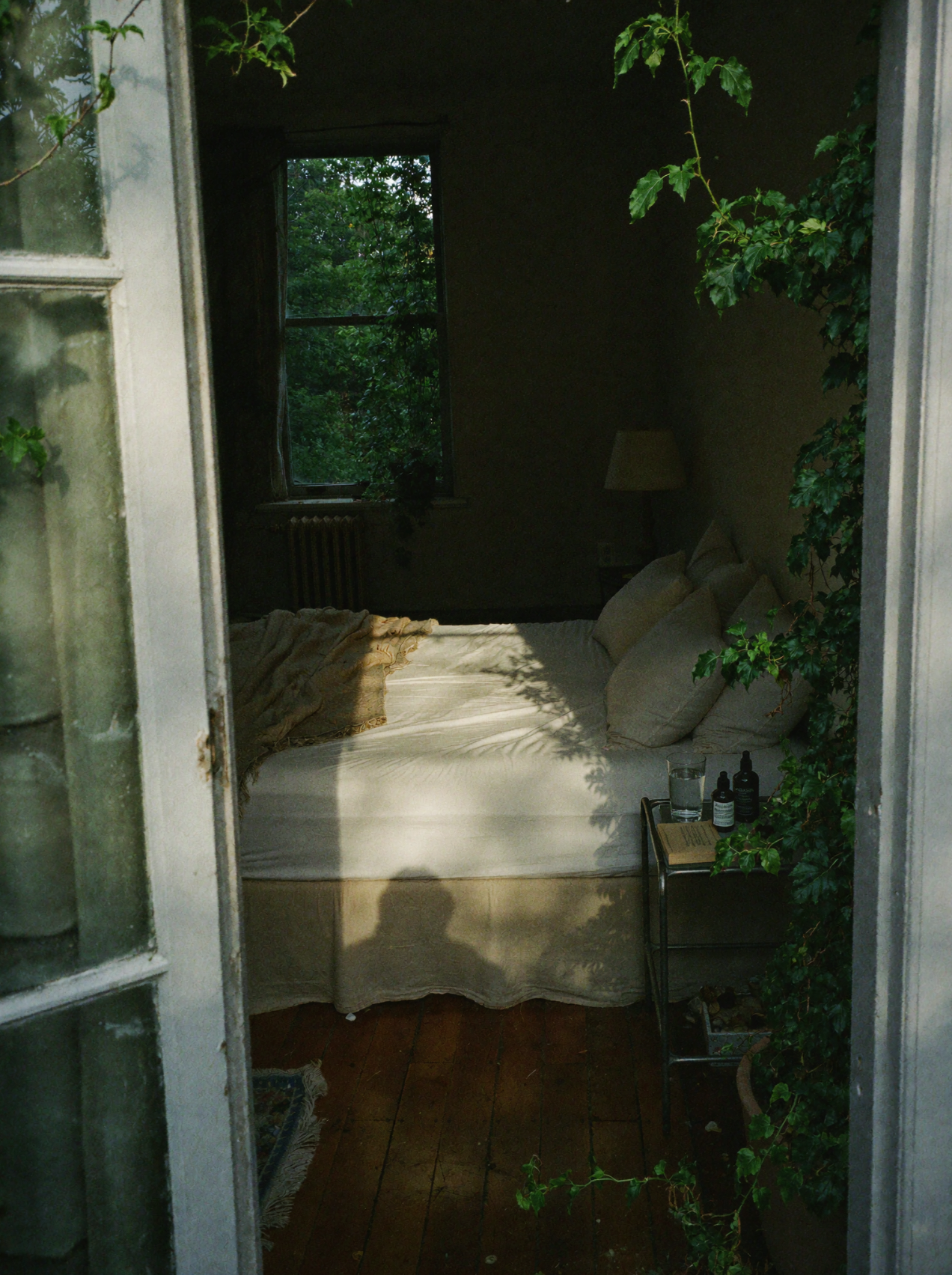 View through an ivy-framed doorway into a sunlit bedroom with skincare bottles on the bedside table