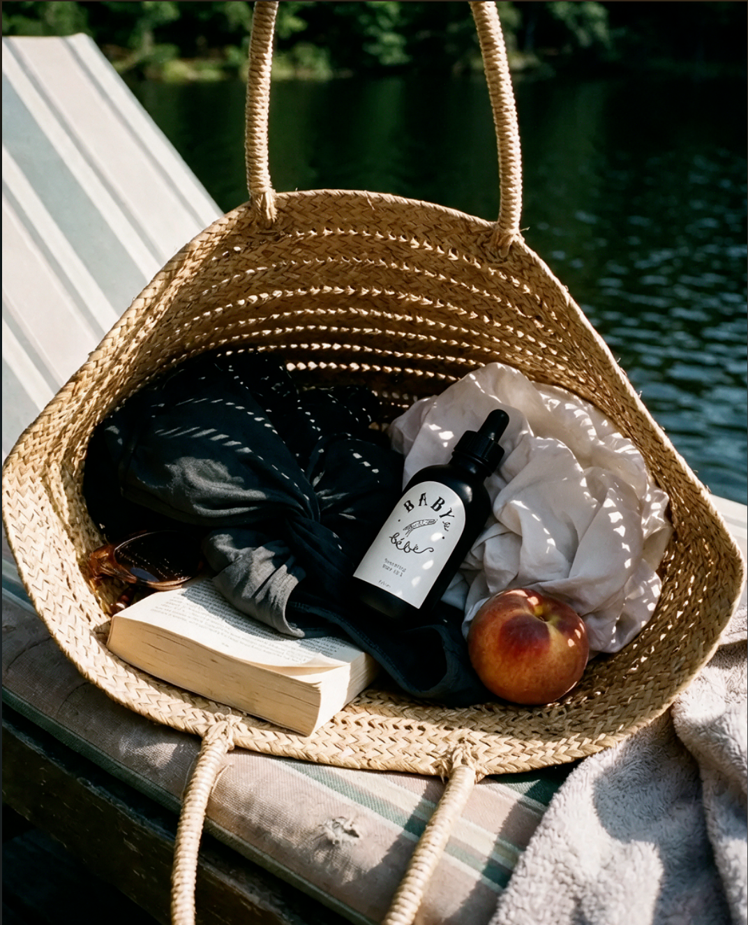 Straw tote by the water holding a Baby le Bébé oil, a book and a peach