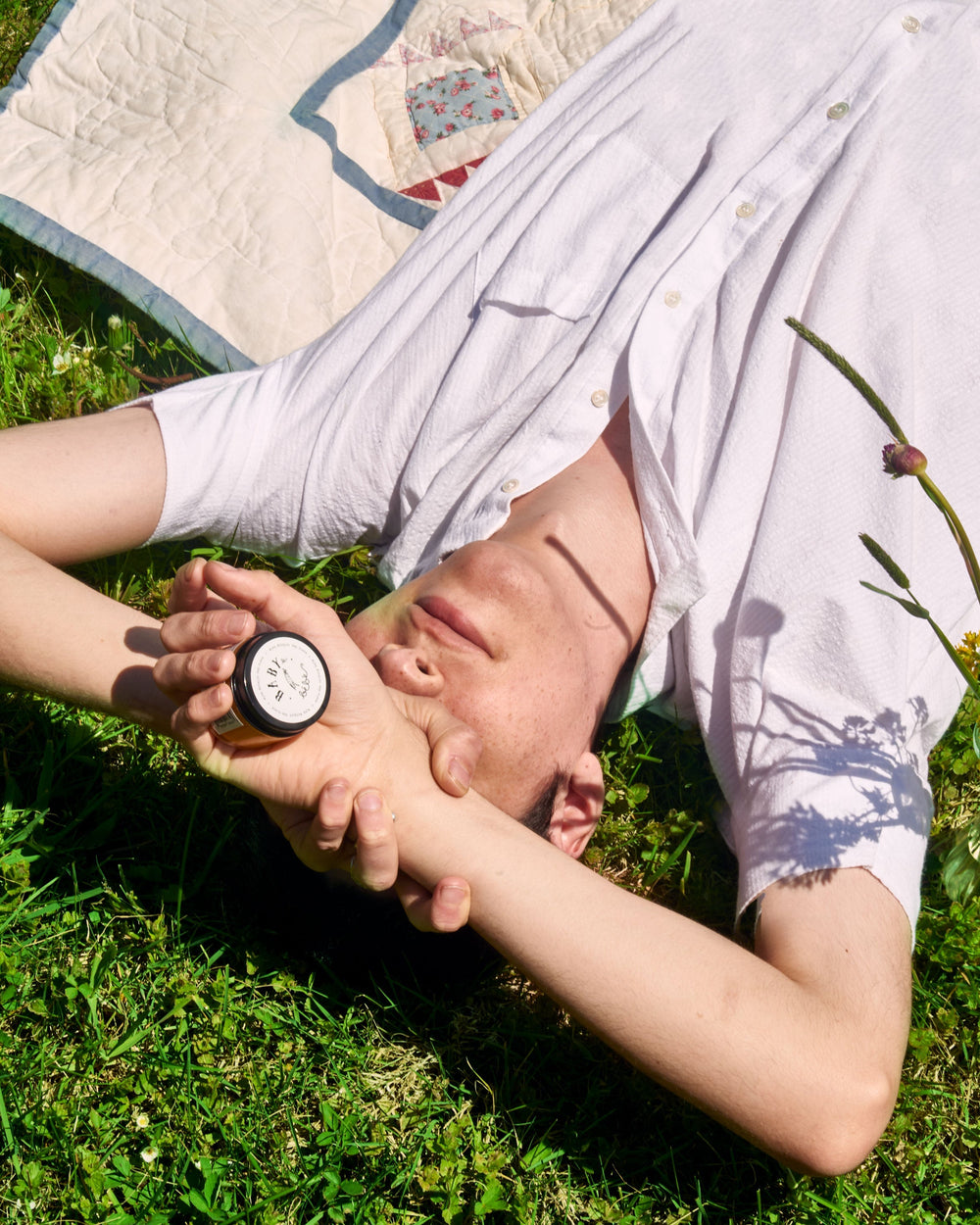 Young woman holding a container of balm over her head and lying in the grass.
