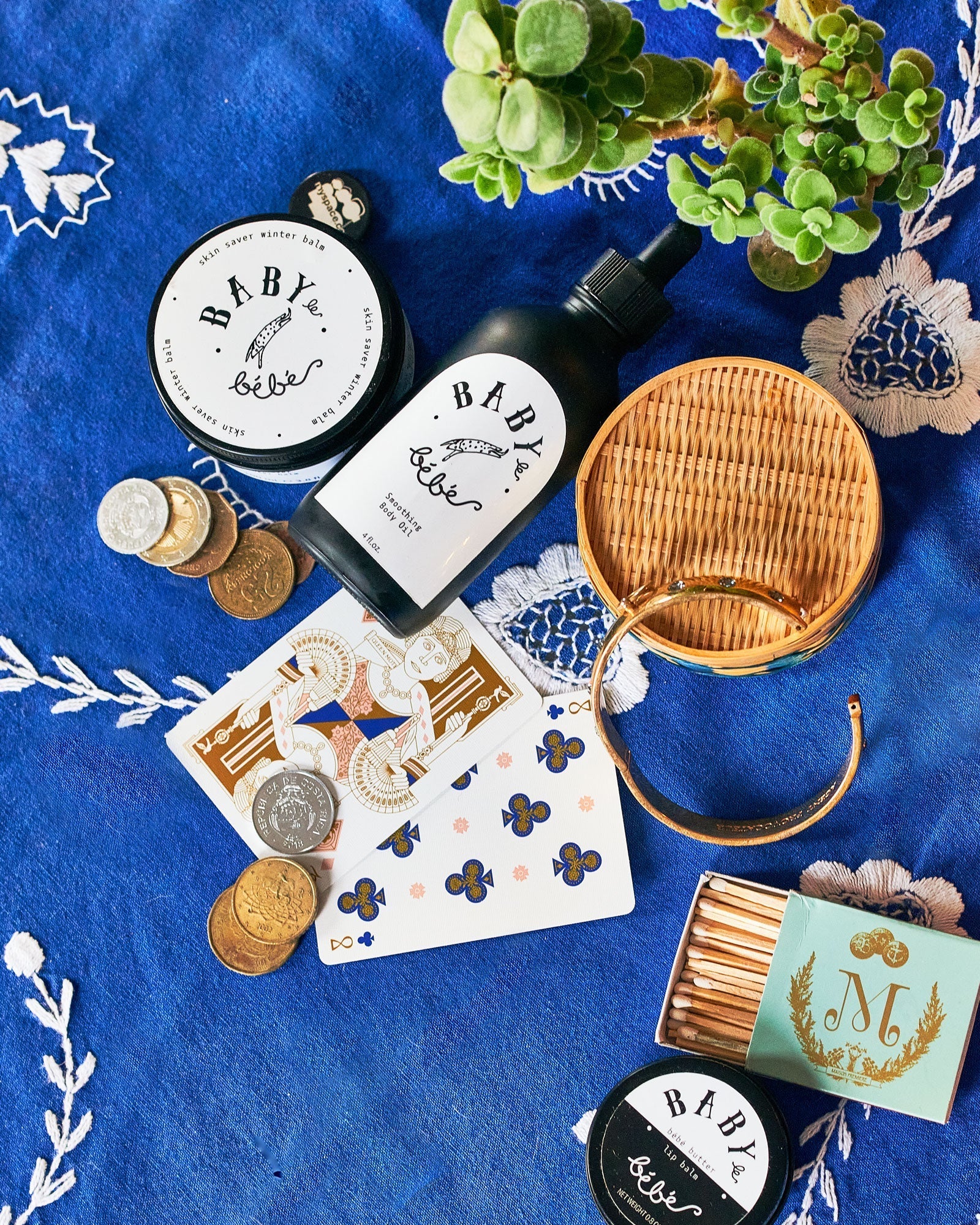 Baby le Bébé tins and a bottle with playing cards, coins and a woven fan on blue embroidered cloth