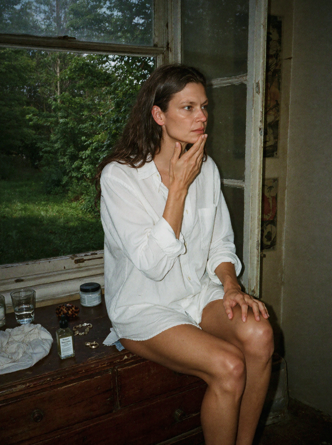 Woman in a white shirt seated by an open window with skincare bottles on the ledge