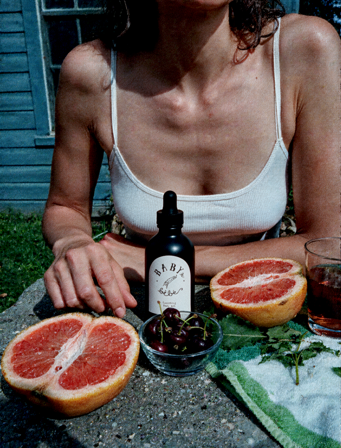 Woman in a white camisole at an outdoor table with a Baby le Bébé dropper bottle, halved grapefruit and cherries