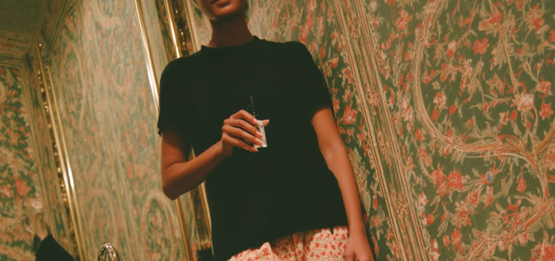 Woman in a black T-shirt holding a small bottle against floral wallpaper