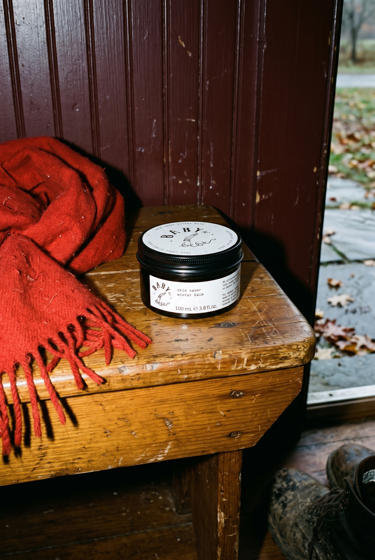 Baby le Bébé Skin Saver Winter Balm on a worn wooden bench beside a red wool scarf, with muddy boots by a door open to autumn leaves.