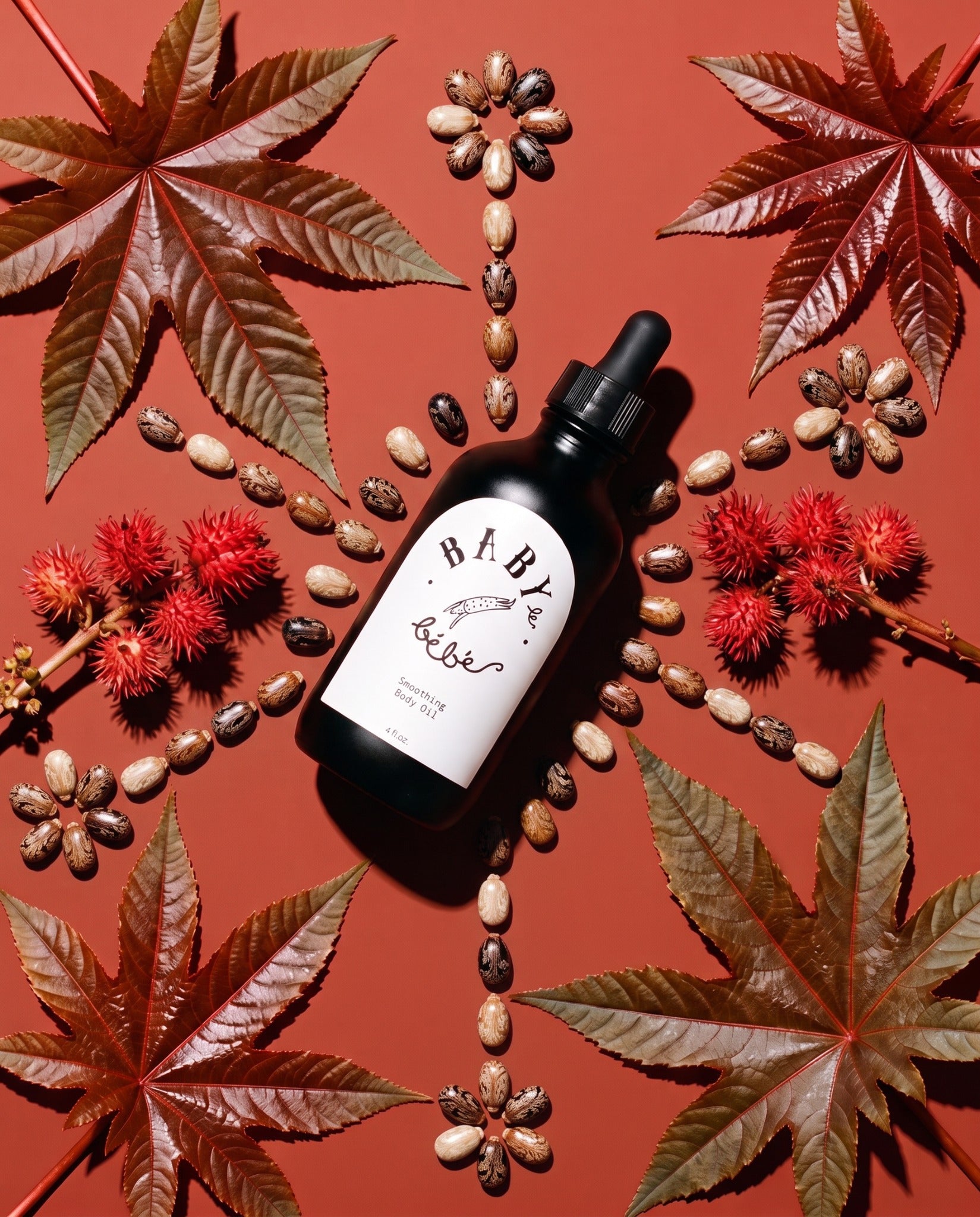Smoothing Body Oil surrounded by castor leaves, mottled seeds and red seed pods on a rust-colored background.